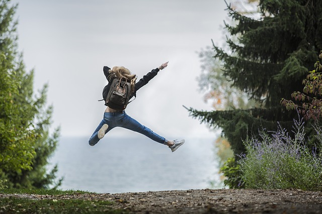 Nuori nainen hyppää ilmaan reppu selässä, taustalla epäselvä merinäköala ja puita.
